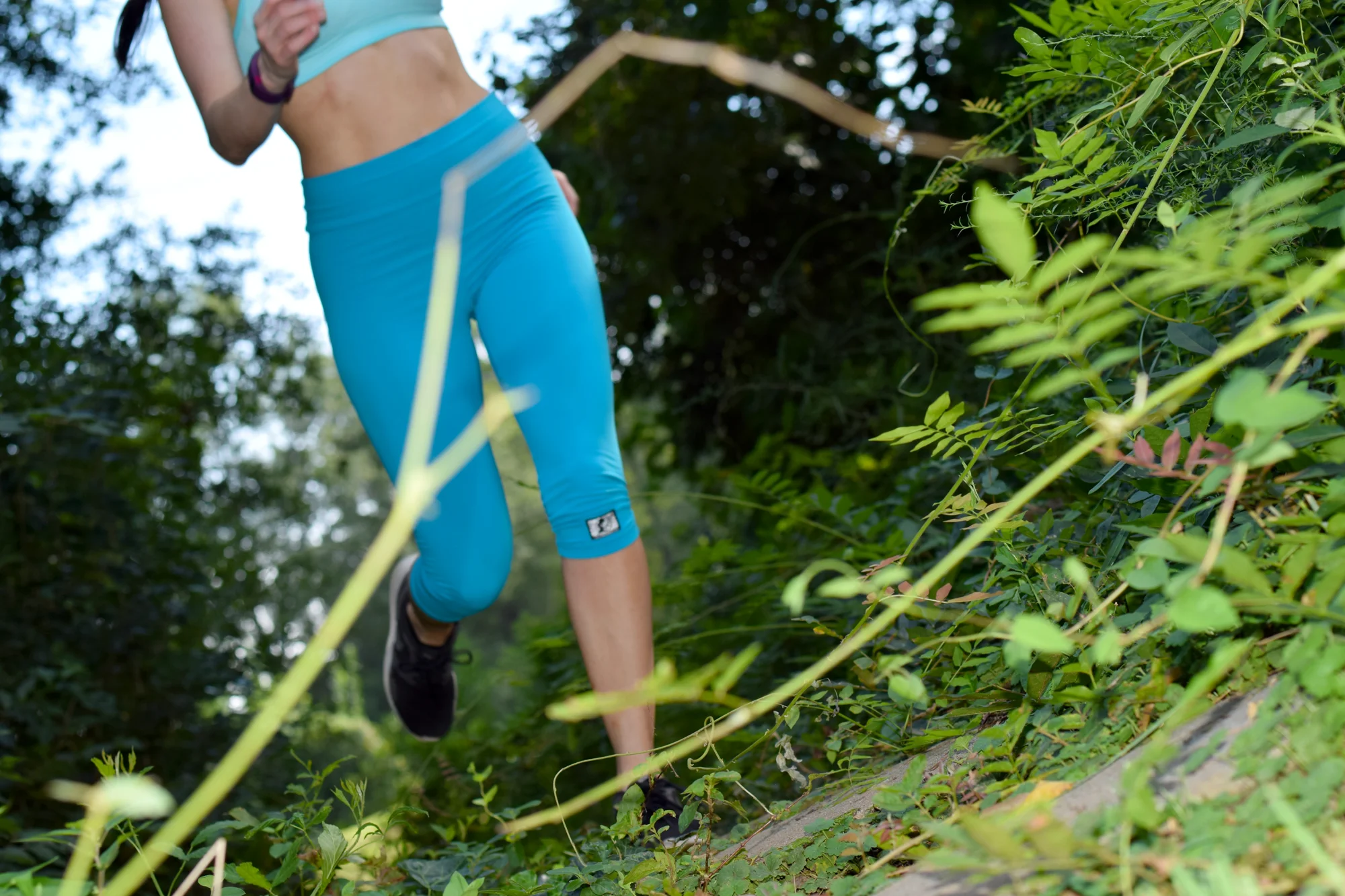 Close-up of a runner's legs in teal leggings moving along a green, leafy trail, with vibrant green and blue tones and an energetic mood
