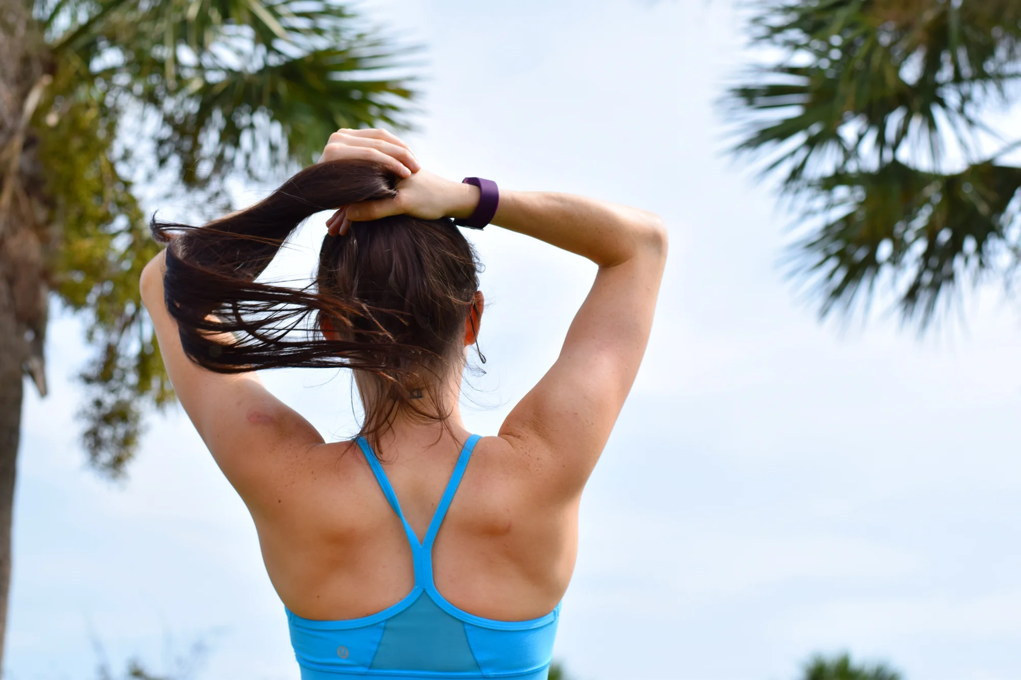 Woman seen from behind putting her hair up in a blue sports top, palm trees against pale sky