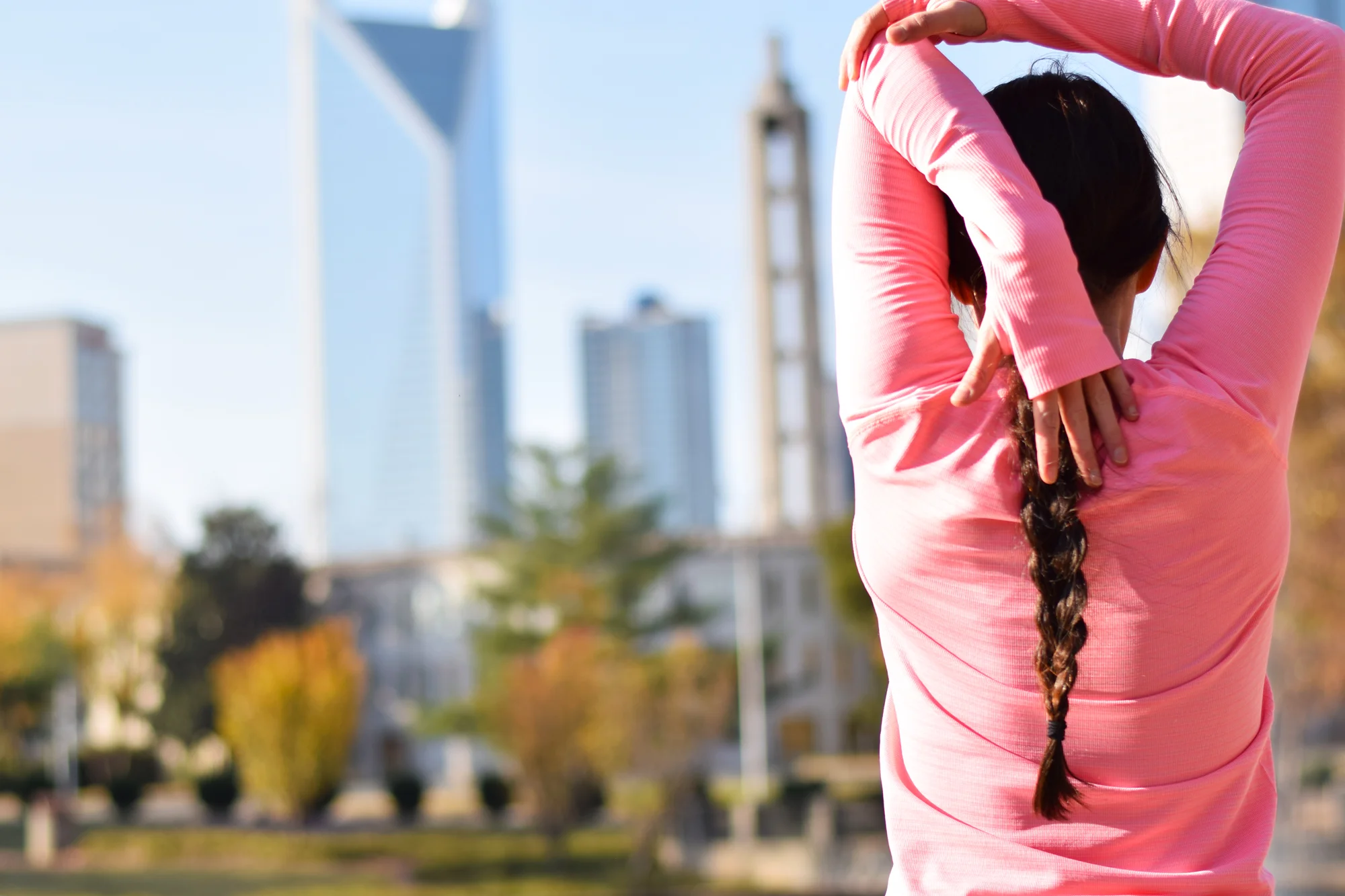 Woman in a pink long-sleeve top stretching with arms overhead, city skyline with towers in the background
