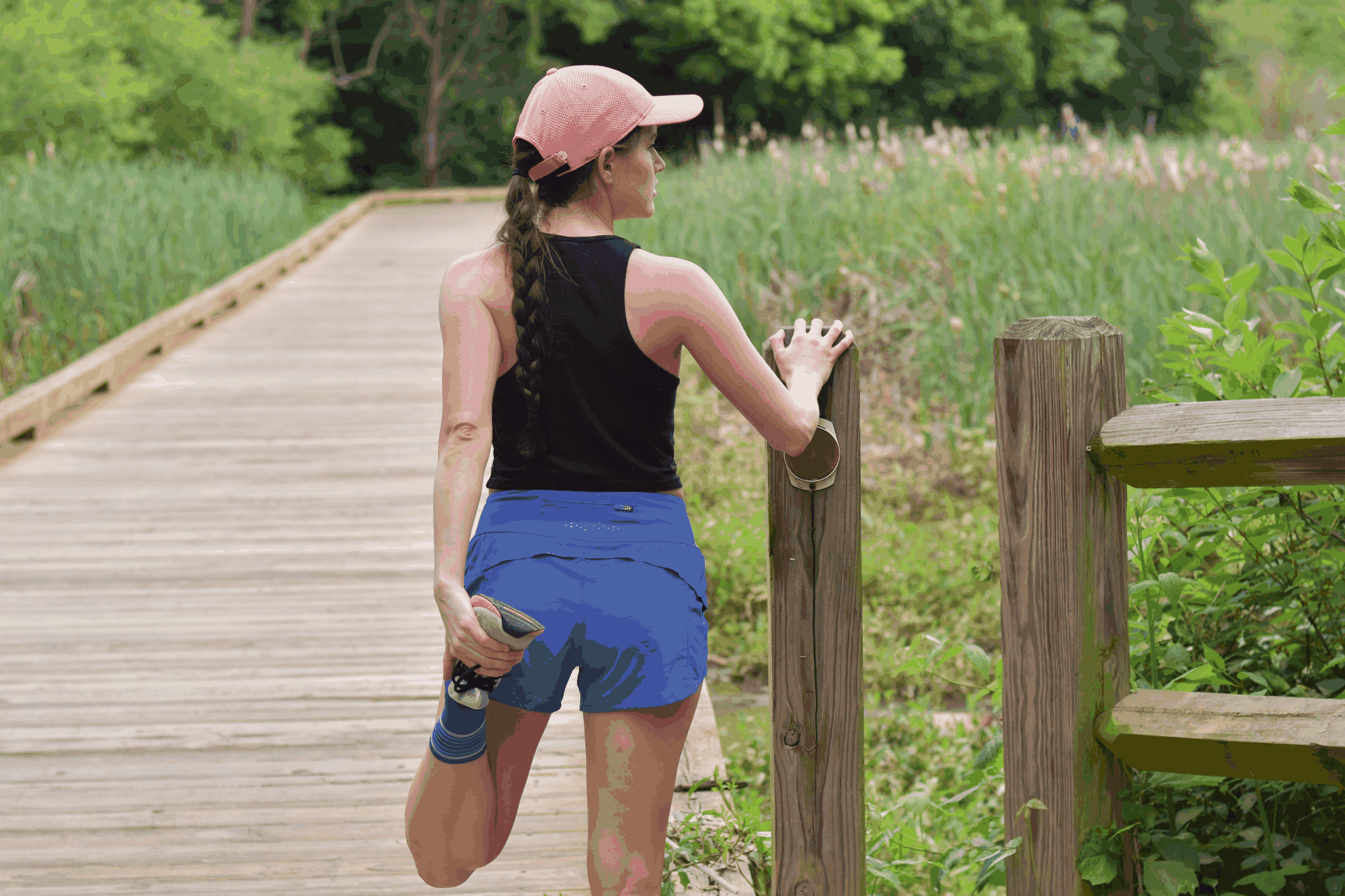 Woman stretching her leg on a wooden boardwalk near tall grasses, wearing a black tank top, calm outdoor scene